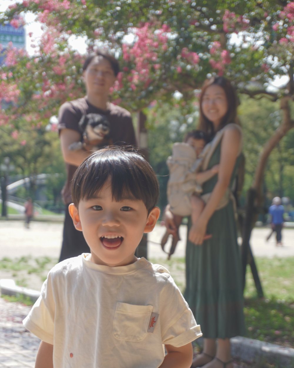 満面の笑みで駆けてくる男の子と、その後ろで微笑む両親、赤ちゃん、愛犬。花の咲く木の下での家族写真